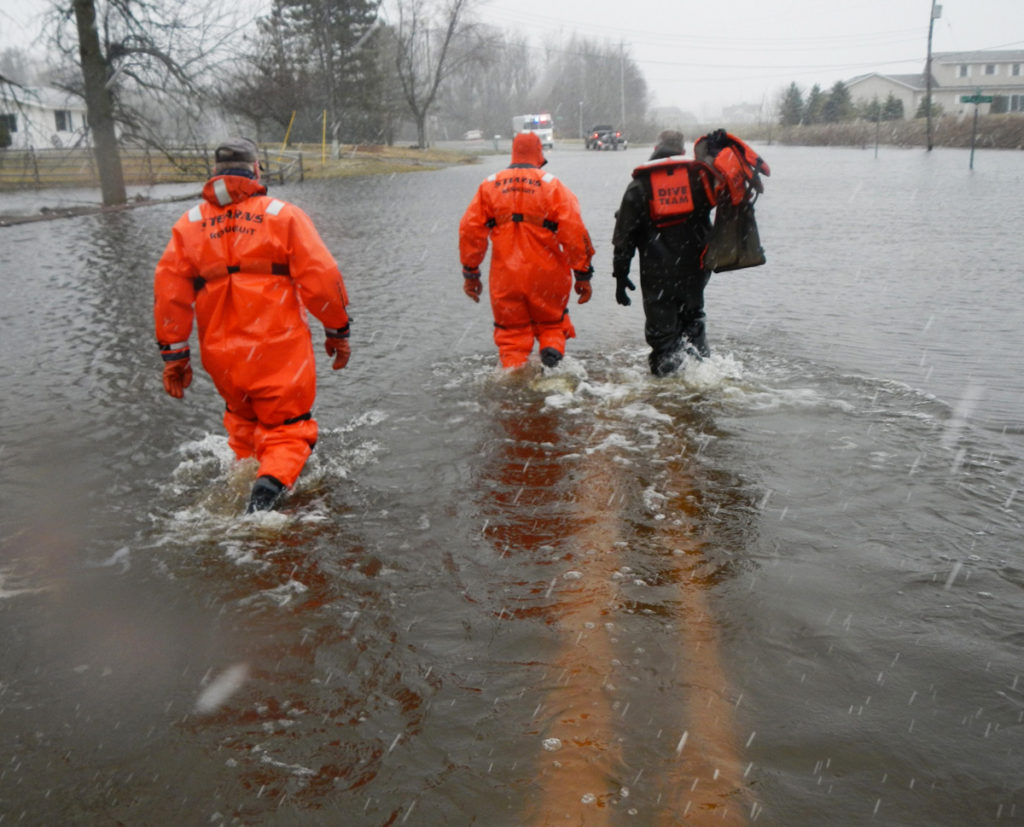 A rescue team in jumpsuits carrying life jackets through the flood waters on a road that is completely underwater.